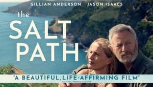 Gillian Anderson and Jason Isaacs as Raynor and Moth Winn in The Salt Path, sitting on a coastal cliffside with the ocean and rugged landscape behind them.
